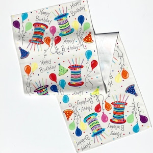 May include: White fabric with a colorful birthday design. The fabric features the words "Happy Birthday" along with illustrations of birthday cakes, party hats, and balloons in various colors. The fabric is folded.