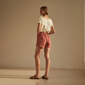 Pode incluir: Uma mulher vestindo uma blusa de linho branca e shorts de linho terracota. Ela está de pé de costas para a câmera com as mãos nos bolsos.
