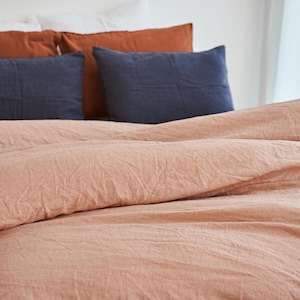 May include: A textured peach-colored duvet cover draped over a bed, accompanied by two dark blue rectangular pillows and two burnt orange square pillows, all featuring a visible woven fabric grain and a soft, slightly wrinkled appearance.