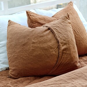 May include: Two rectangular pillowcases in a solid rust-brown color with a visible overlapping envelope closure on the front. The pillowcases feature a textured fabric surface and are positioned alongside a white pillowcase on a bed.