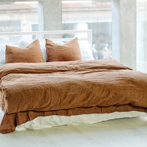 May include: A bed featuring a rust-colored duvet cover and two matching pillowcases, layered over a white bed skirt and white pillows. The bedding set has a visible textured fabric finish and is arranged on a white frame in a bright room.