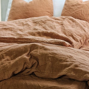 May include: A bedding set featuring a duvet cover and two pillowcases in a solid, earthy terracotta brown color. The fabric has a visible, textured weave with a soft, rumpled appearance, creating a layered look across the bed surface.