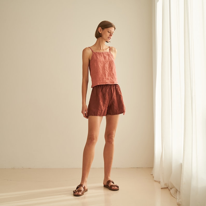 Pode incluir: Uma mulher vestindo um top de tanque de linho cor de ferrugem e shorts de linho cor de ferrugem combinando. Ela est&aacute; em p&eacute; na frente de uma parede branca e uma cortina branca.