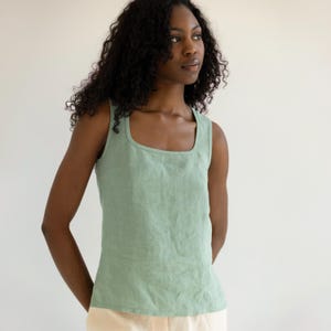 May include: A sage green sleeveless tank top featuring a square neckline and a fitted silhouette. The garment is constructed from a textured fabric with visible seams along the bodice, providing a structured look with wide shoulder straps.