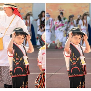 May include: A young boy wearing a black vest with colorful embroidery, a white shirt, and a black hat. He is holding the hat with both hands and looking at the camera.