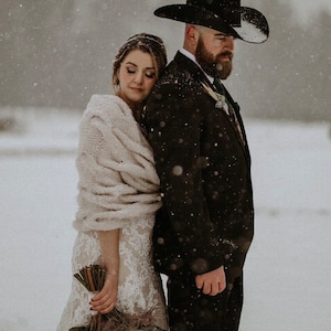 Op de afbeelding: Een bruid en bruidegom staan dicht bij elkaar in een besneeuwd landschap. De bruid draagt een witte kanten trouwjurk en een cr&egrave;mekleurige sjaal. De bruidegom draagt een zwart pak en een cowboyhoed. De bruid houdt een boeket bloemen vast.