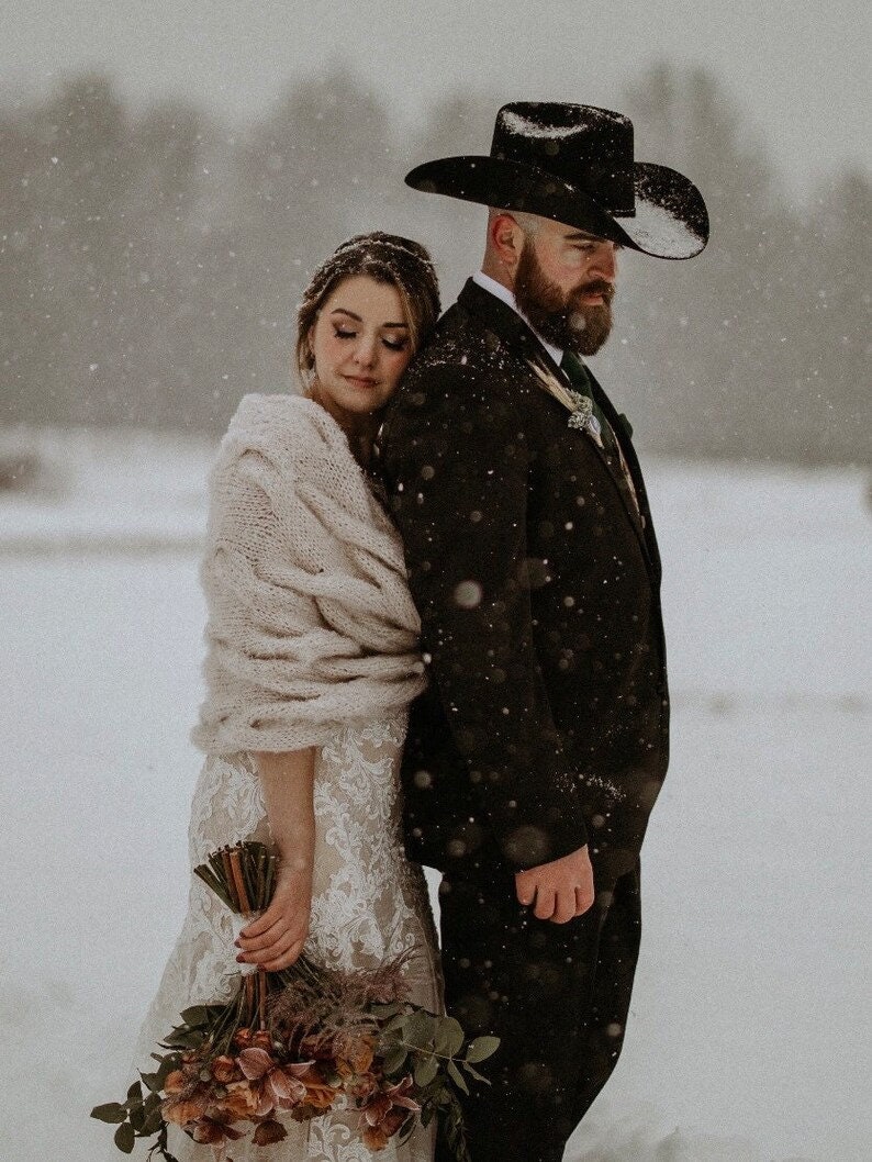 Op de afbeelding: Een bruid en bruidegom staan dicht bij elkaar in een besneeuwd landschap. De bruid draagt een witte kanten trouwjurk en een cr&egrave;mekleurige sjaal. De bruidegom draagt een zwart pak en een cowboyhoed. De bruid houdt een boeket bloemen vast.