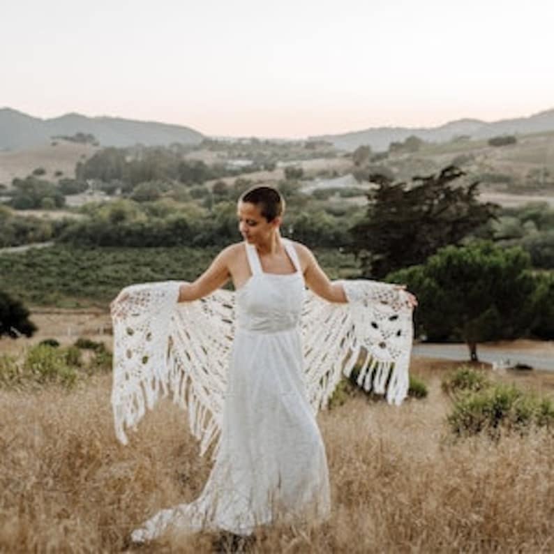 May include: A white crochet shawl with fringe, worn over a white dress. The person is standing in a field with hills in the background.