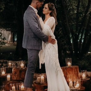 May include: A couple in a romantic embrace, the bride wearing a white lace dress and the groom in a blue suit. They are surrounded by candlelight in a rustic outdoor setting.