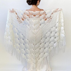 May include: White crochet shawl with a fringe border and a delicate lace pattern. The shawl is made with a circular design and has a scalloped edge.