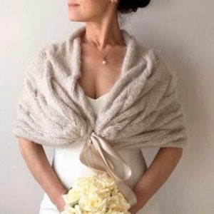 Op de afbeelding: Een beige gebreide sjaal met een kabelpatroon. De sjaal is gedrapeerd over een witte jurk en vastgemaakt met een satijnen lint. De persoon houdt een boeket witte bloemen vast. Een parelketting is zichtbaar.