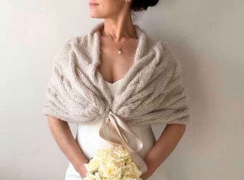 Op de afbeelding: Een beige gebreide sjaal met een kabelpatroon. De sjaal is gedrapeerd over een witte jurk en vastgemaakt met een satijnen lint. De persoon houdt een boeket witte bloemen vast. Een parelketting is zichtbaar.