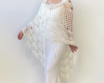 Könnte beinhalten: Ein cremefarbener, offen gewebter Häkelschal mit einem Muscheldesign und Fransenbesatz. Der Schal ist über einem weißen Kleid drapiert und erzeugt einen fließenden, eleganten Look. Das komplizierte Muster und die Textur des Häkels sind deutlich sichtbar.