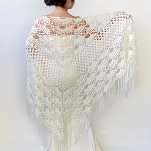 May include: White crochet shawl with a delicate lace pattern and fringe trim. The shawl is made of a lightweight yarn and has a scalloped edge.
