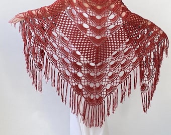 May include: A rust-colored crocheted shawl with a fringe border. The shawl has a delicate openwork pattern and is made with a soft yarn.