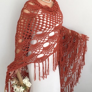 May include: A terracotta crocheted shawl with fringe. The shawl is draped over a white dress and has a delicate, openwork design.