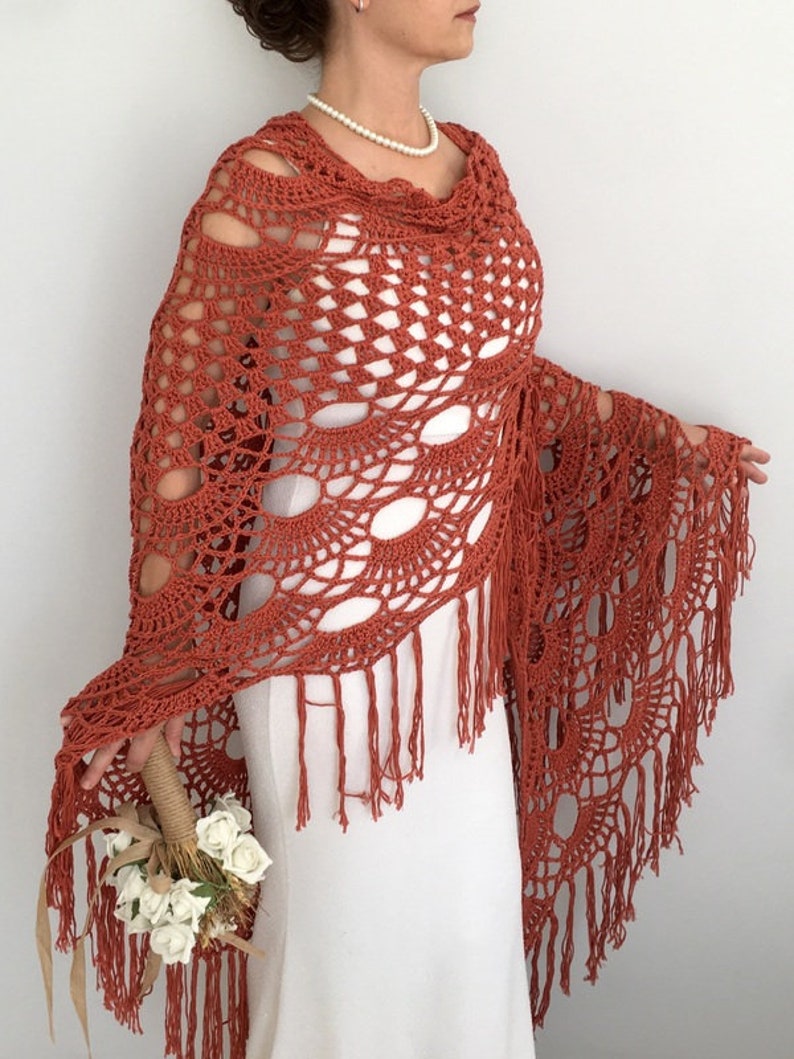 May include: A terracotta crocheted shawl with fringe. The shawl is draped over a white dress and has a delicate, openwork design.
