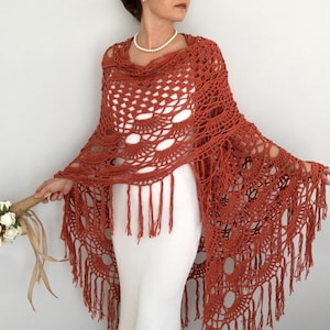 May include: A terracotta crocheted shawl with fringe. The shawl has a delicate openwork design and is draped over a white dress.