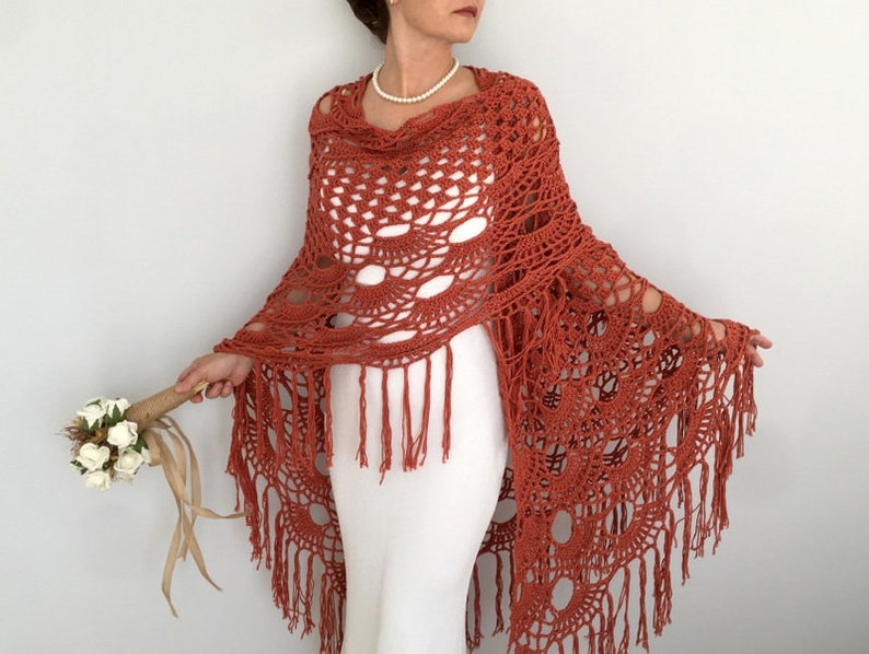 May include: A terracotta crocheted shawl with fringe. The shawl has a delicate openwork design and is draped over a white dress.