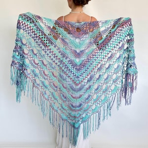 May include: A handcrafted, open-weave shawl in teal, purple, and lavender hues. The shawl showcases a geometric pattern with scalloped edges and long fringe. The crochet design is displayed against a white backdrop.