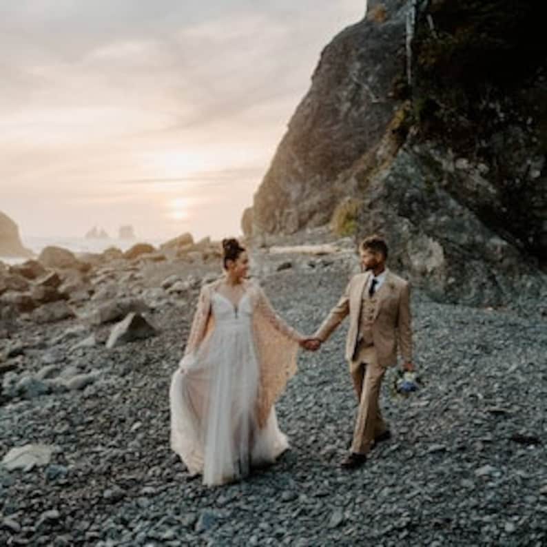 May include: A couple walks hand-in-hand on a rocky beach at sunset. The bride wears a flowing white gown with a lace overlay, while the groom wears a tan suit. The couple is smiling and looking at each other.