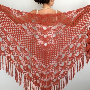 May include: A crocheted shawl in a burnt orange color with a fringe border. The shawl has a delicate openwork pattern with large oval shapes.