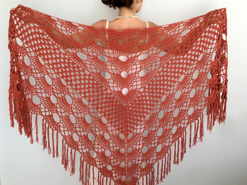 May include: A crocheted shawl in a burnt orange color with a fringe border. The shawl has a delicate openwork pattern with large oval shapes.