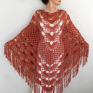 May include: A terracotta crocheted shawl with a delicate openwork pattern and fringe trim. The shawl is draped over a person's shoulders.