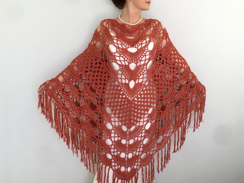 May include: A terracotta crocheted shawl with a delicate openwork pattern and fringe trim. The shawl is draped over a person's shoulders.