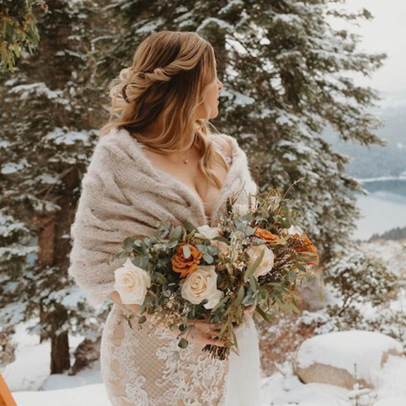 Op de afbeelding: Een bruid in een winterse setting, met een cr&egrave;mekleurige sjaal en een boeket bloemen. Het boeket bevat witte en oranje rozen, met groen. De achtergrond toont besneeuwde bomen en een meer.