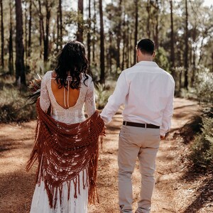 May include: A couple walks hand-in-hand down a dirt path in a forest. The woman is wearing a white lace wedding dress with a brown crocheted shawl. The man is wearing a white button-down shirt and beige pants.