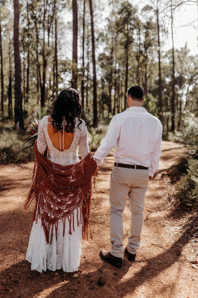 May include: A couple walks hand-in-hand down a dirt path in a forest. The woman is wearing a white lace wedding dress with a brown crocheted shawl. The man is wearing a white button-down shirt and beige pants.