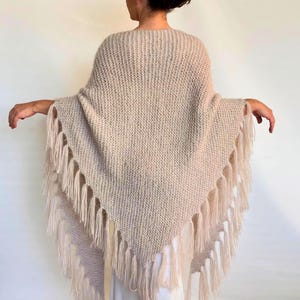 Puede incluir: Un poncho tejido de color beige con un dobladillo con flecos. El poncho está hecho de un hilo suave y texturizado y tiene forma triangular. La franja es larga y cuelga del borde inferior del poncho.