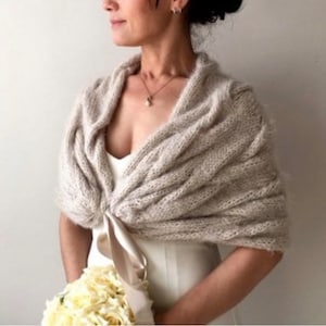 Op de afbeelding: Beige gebreide sjaal met een gedrapeerd ontwerp. De sjaal is vastgebonden met een satijnen lint. Het model draagt een witte jurk en houdt een boeket witte bloemen vast. De sjaal is een bruiloftsaccessoire.