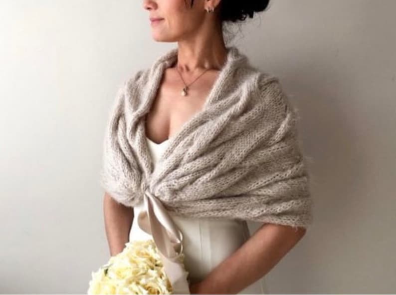 Op de afbeelding: Beige gebreide sjaal met een gedrapeerd ontwerp. De sjaal is vastgebonden met een satijnen lint. Het model draagt een witte jurk en houdt een boeket witte bloemen vast. De sjaal is een bruiloftsaccessoire.