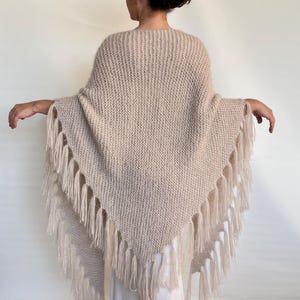 Puede incluir: Un poncho tejido de color beige con flecos. El poncho tiene forma de triángulo y presenta un patrón de tejido texturizado. Los flecos son de un tono beige más claro y cuelgan del borde inferior del poncho.