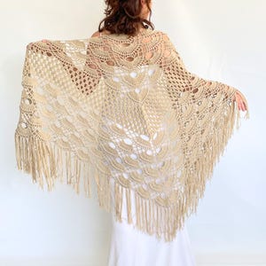 Peut inclure: Un châle en crochet beige avec un motif ajouré détaillé et une bordure à franges. Le châle est drapé sur les épaules, mettant en valeur les motifs et la texture complexes. Le châle est de couleur beige clair.