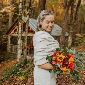 Op de afbeelding: Een vrouw in een witte jurk en een cr&egrave;mekleurige imitatiebont sjaal houdt een levendig boeket zonnebloemen, rode bloemen en groen vast. De achtergrond toont een kleine houten constructie met een golfplaten dak, omringd door bomen.