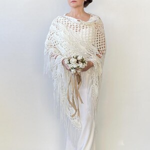 May include: White crochet shawl with fringe, worn over a white dress. The person is holding a small bouquet of white flowers.