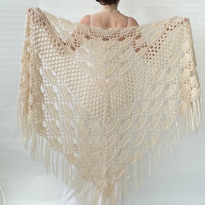 May include: Cream-colored, open-weave shawl with a triangular shape and fringe along the edges. The shawl features a detailed crochet pattern with scalloped and geometric designs. The delicate fabric is held up, showcasing its intricate craftsmanship.