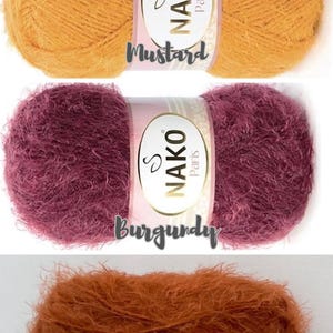 Op de afbeelding: Drie bollen Nako Paris garen in mosterd, bordeaux en kaneel kleuren. Elke bol heeft een label met de merknaam en kleurnaam. Het garen ziet er zacht en pluizig uit, geschikt voor brei- of haakprojecten.