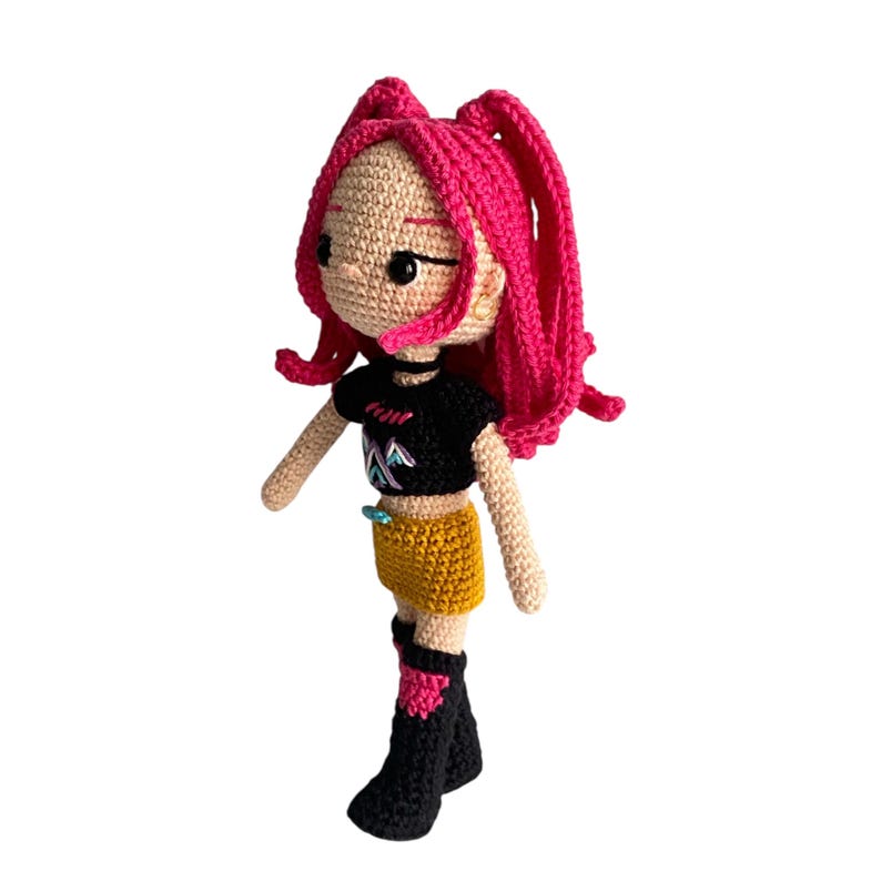 May include: Crocheted doll with pink hair styled in two ponytails. The doll wears a black t-shirt, mustard yellow shorts, and black boots with pink accents. The doll has a light skin tone and black eyes.