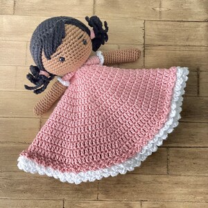 以下が含まれることがあります： 茶色の肌と黒髪のおさげをした、ピンクのレースの縁取りのあるピンクのドレスを着た、かぎ針編みのドールです。