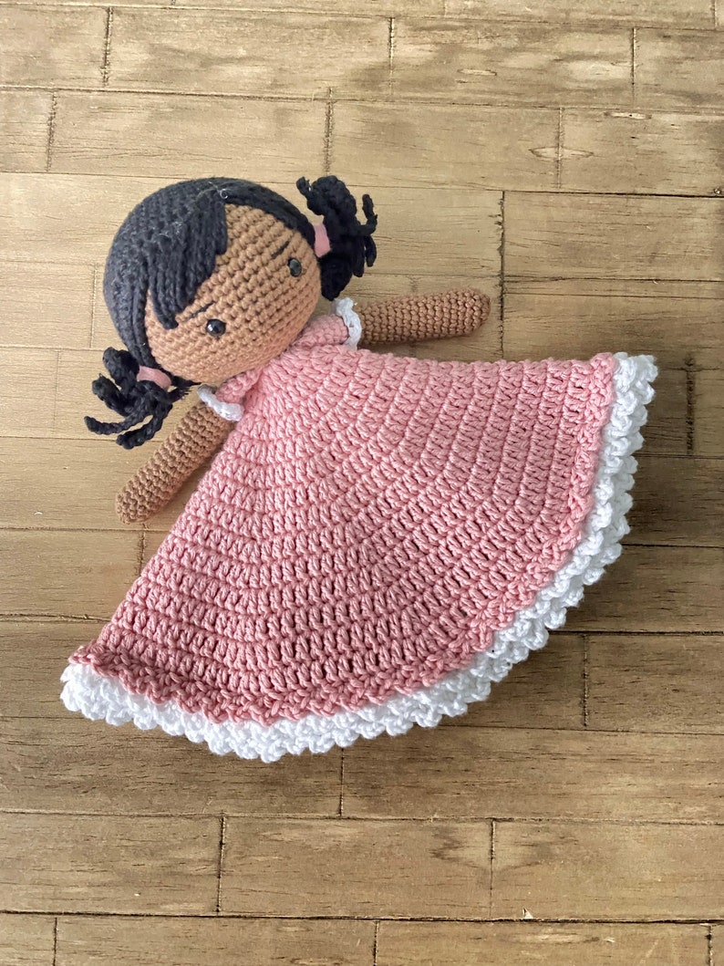 以下が含まれることがあります： 茶色の肌と黒髪のおさげをした、ピンクのレースの縁取りのあるピンクのドレスを着た、かぎ針編みのドールです。