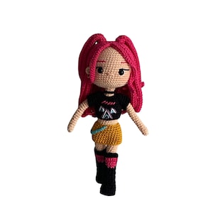 May include: A handmade crochet doll with pink hair styled in two high ponytails. The doll wears a black top with a graphic design, a yellow skirt, and black boots with pink accents. The doll has a tan skin tone and is set against a white background.