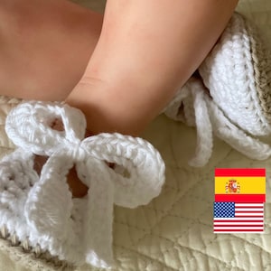 May include: A pair of handmade crochet baby booties featuring a tan base and a white upper section. Each bootie is adorned with a prominent white crocheted bow tied at the ankle. The design includes a visible Spanish flag and an American flag icon.