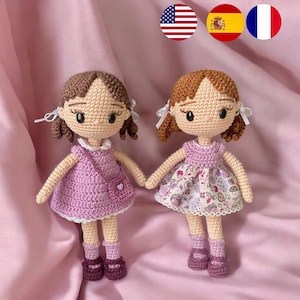Puede incluir: Dos muñecas de crochet con pelo castaño y vestidos rosas. Una muñeca lleva un vestido morado con ribete de encaje blanco y un pequeño bolsillo. La otra muñeca lleva un vestido floral con ribete de encaje blanco.