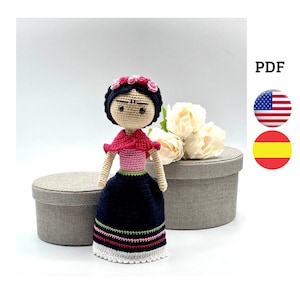May include: Crochet doll pattern in English and Spanish. The doll is wearing a black dress with pink and green stripes, a pink top, and a red shawl. She has black hair and a flower crown.