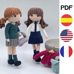 Puede incluir: Dos muñecas de crochet vestidas con uniformes escolares. Una muñeca lleva un suéter verde y una falda gris, mientras que la otra muñeca lleva una camisa blanca y una falda azul. Ambas muñecas tienen cabello castaño y llevan bolsos. Las muñecas están de pie sobre una superficie blanca. La imagen también incluye una chaqueta de crochet roja y una bolsa de crochet azul.