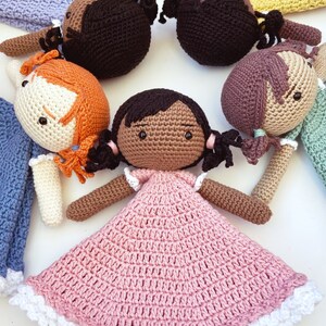 以下が含まれることがあります： 異なる色の髪と服を着た、かぎ針編みの人形がいくつかあります。人形は白い表面に円形に配置されています。1体の人形は、白いレースのトリムが付いたピンクのドレスを着ています。